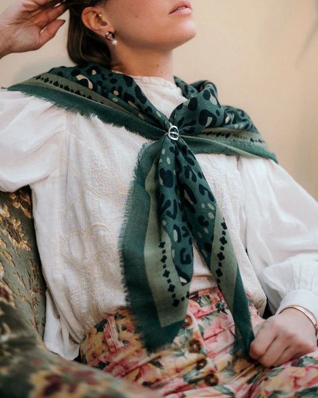 A woman in a white blouse, floral high-waisted pants, and a Maison Lecomte Flament Leo Scarf Green draped around her shoulders sits on an ornate chair, embodying elegance. Her face is partially out of frame.