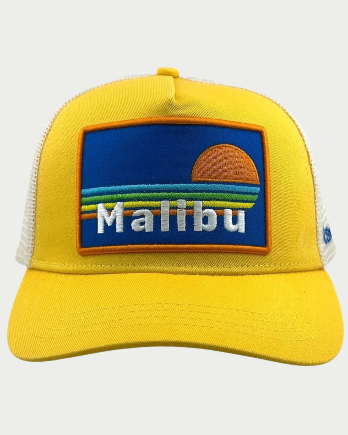 The Soulbyrd Malibu Trucker Hat is yellow and white with a mesh back, featuring a chenille patch of an orange sun, blue and green wave stripes, and "Malibu" in white letters.