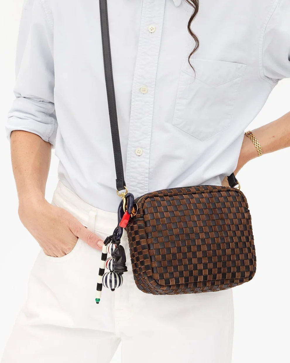 A person in a light blue shirt and white pants holds a brown woven crossbody bag with a black strap, accessorized with the CLARE V. Beaded Stripe Fob Bag Charm. Their hand is in their pocket.