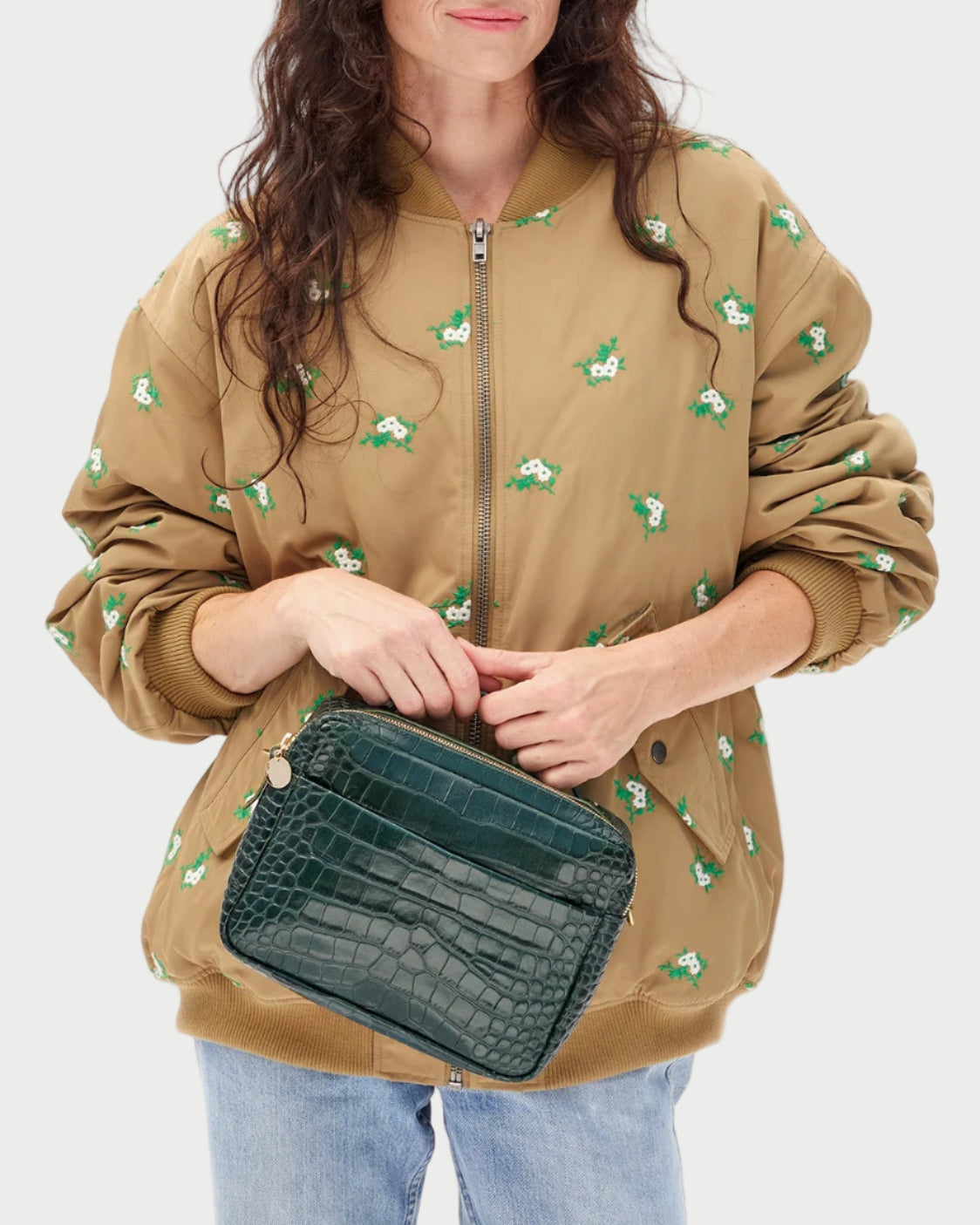 Woman wearing a beige jacket with green patterns, holding a green clutch against a white background