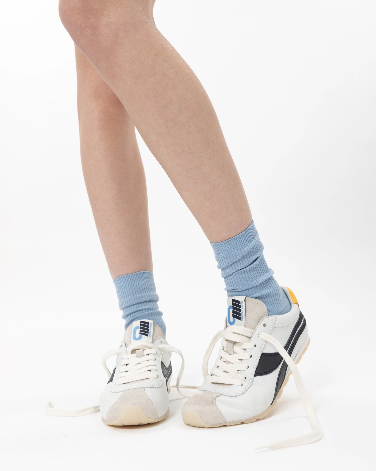 Close-up of a person's legs wearing light blue socks and Oncept Essex Sneaker - Retro Indigo by Oncept, featuring white lightweight design with navy accents and untied laces. Set against a plain white background.