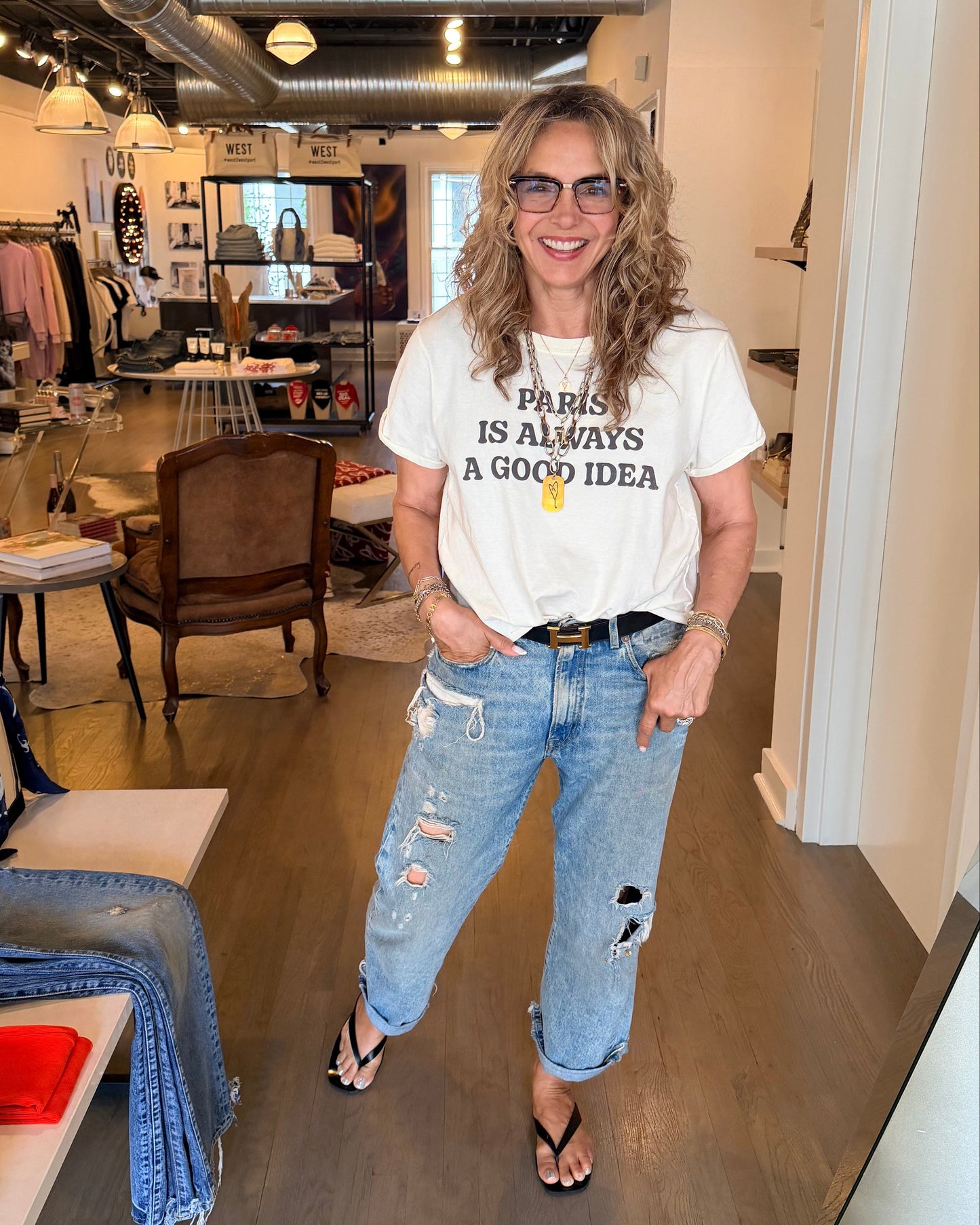 A woman with wavy hair and glasses stands smiling in a stylish boutique, wearing the RECYCLED KARMA Recycled Karma Paris Is Always A Good Idea Tee, paired with ripped jeans and flip-flops. Clothes, chairs, and shelves fill the background.