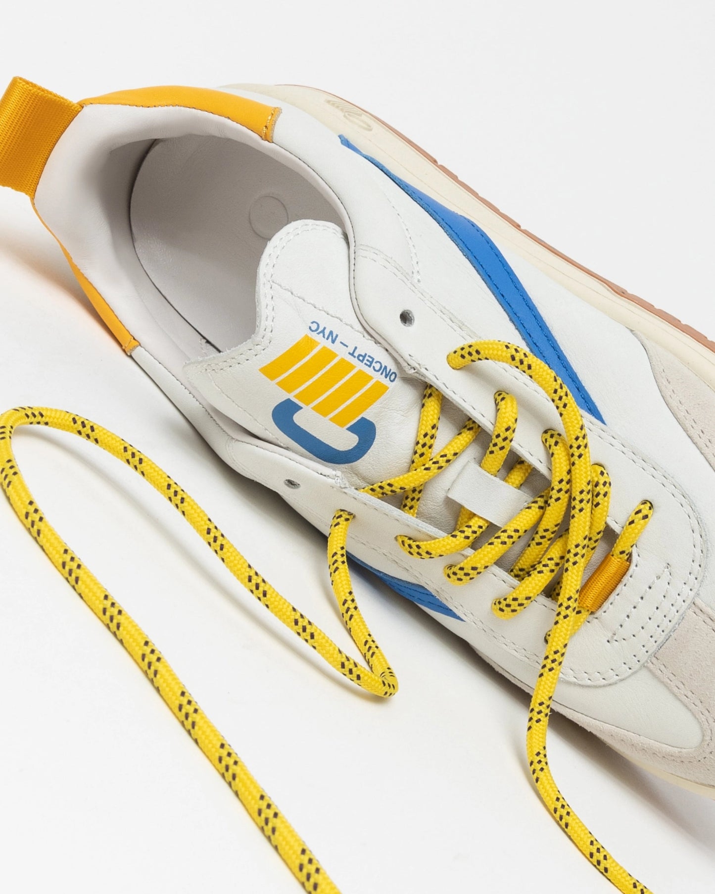 A close-up of the Panama Sneaker by Oncept highlights its comfortable design, showcasing a white base accented with blue and yellow details. The yellow laces are adorned with black spots, and the tongue displays text alongside a stylized graphic. The lace is elegantly arranged in a wavy pattern.