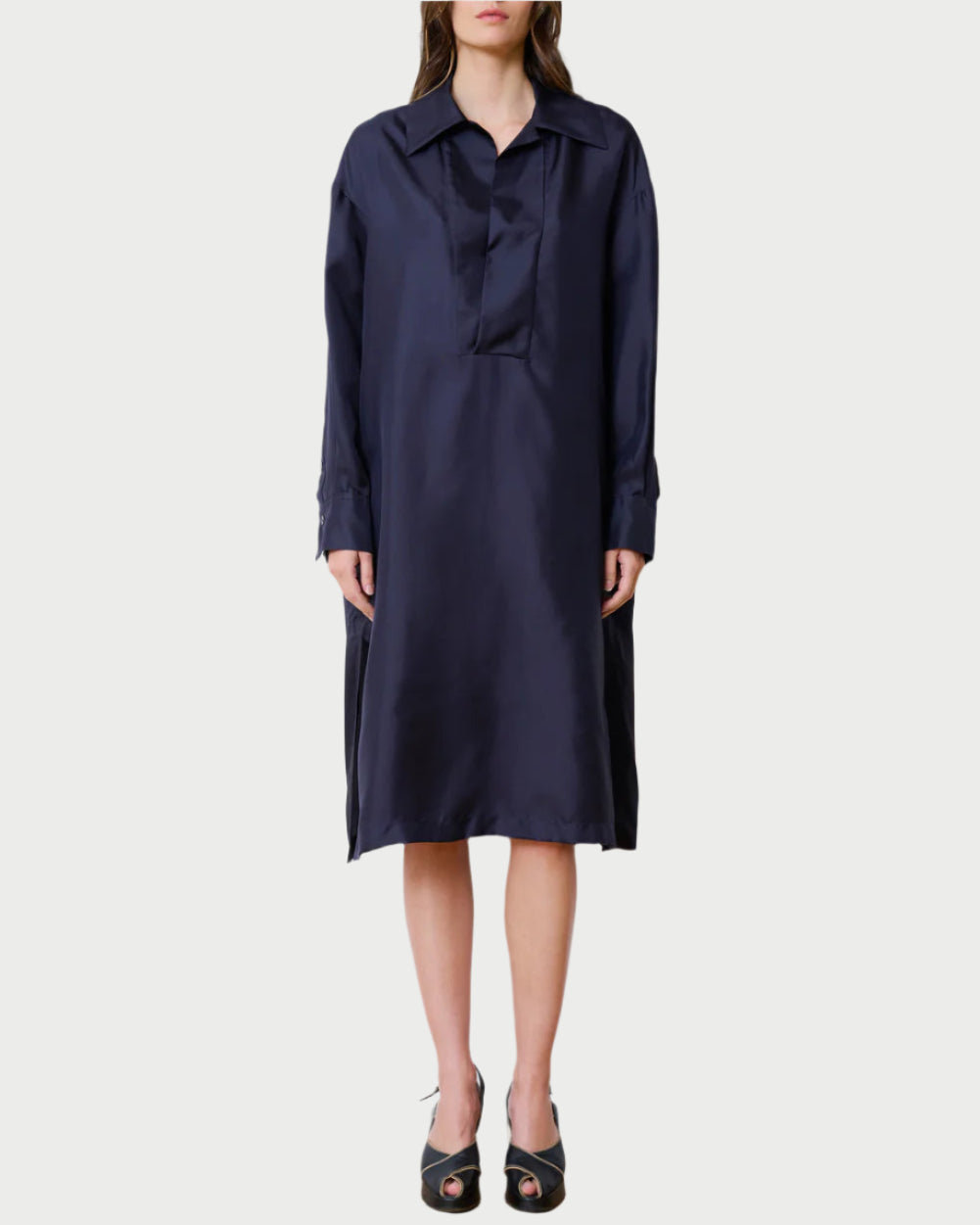 A person dons the Society Dress by WE-AR4, a long-sleeved, dark blue satin shirtdress with a collar and loose-fitting silhouette like a caftan. They pair it with black open-toed heels on a plain white background.