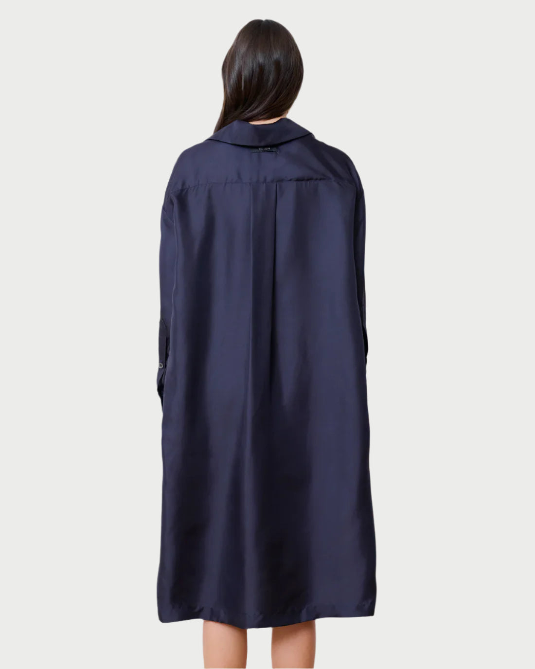 A person with long dark hair stands with their back to the camera, draped in a dark blue Society Dress by WE-AR4. The dress's smooth texture falls elegantly below the knees, exuding sophistication against a plain, off-white background.