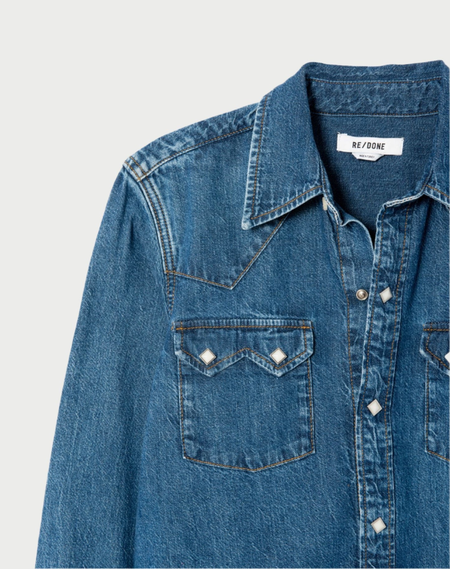 The 50s Western Shirt by RE/DONE offers a deep denim blue hue and showcases a classic pointed collar with snap-button closure, embodying the Western shirt style. It boasts two chest pockets adorned with diamond-shaped snaps and features the "RE/DONE" label inside the collar, making it ideal for those embracing the denim-on-denim trend. Displayed against a light background.