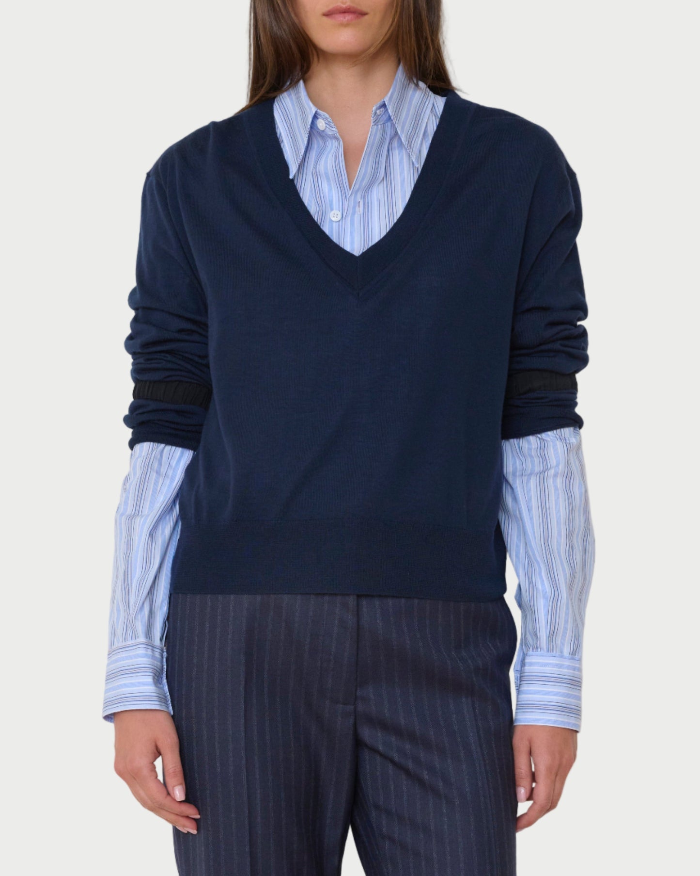 Person wearing a navy sweater over a light blue shirt with dark pinstripe pants against a plain background