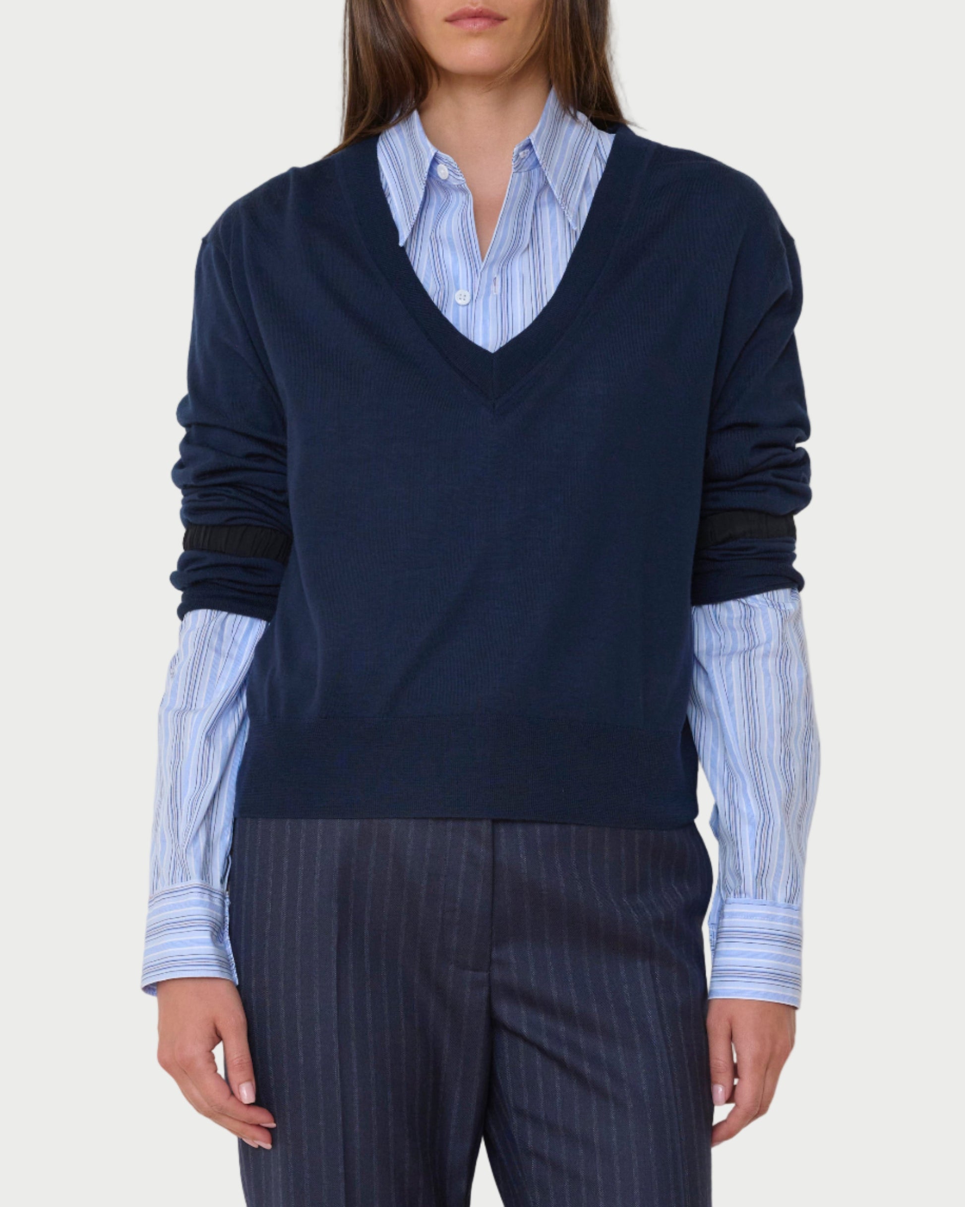 Person wearing a navy sweater over a light blue shirt with dark pinstripe pants against a plain background
