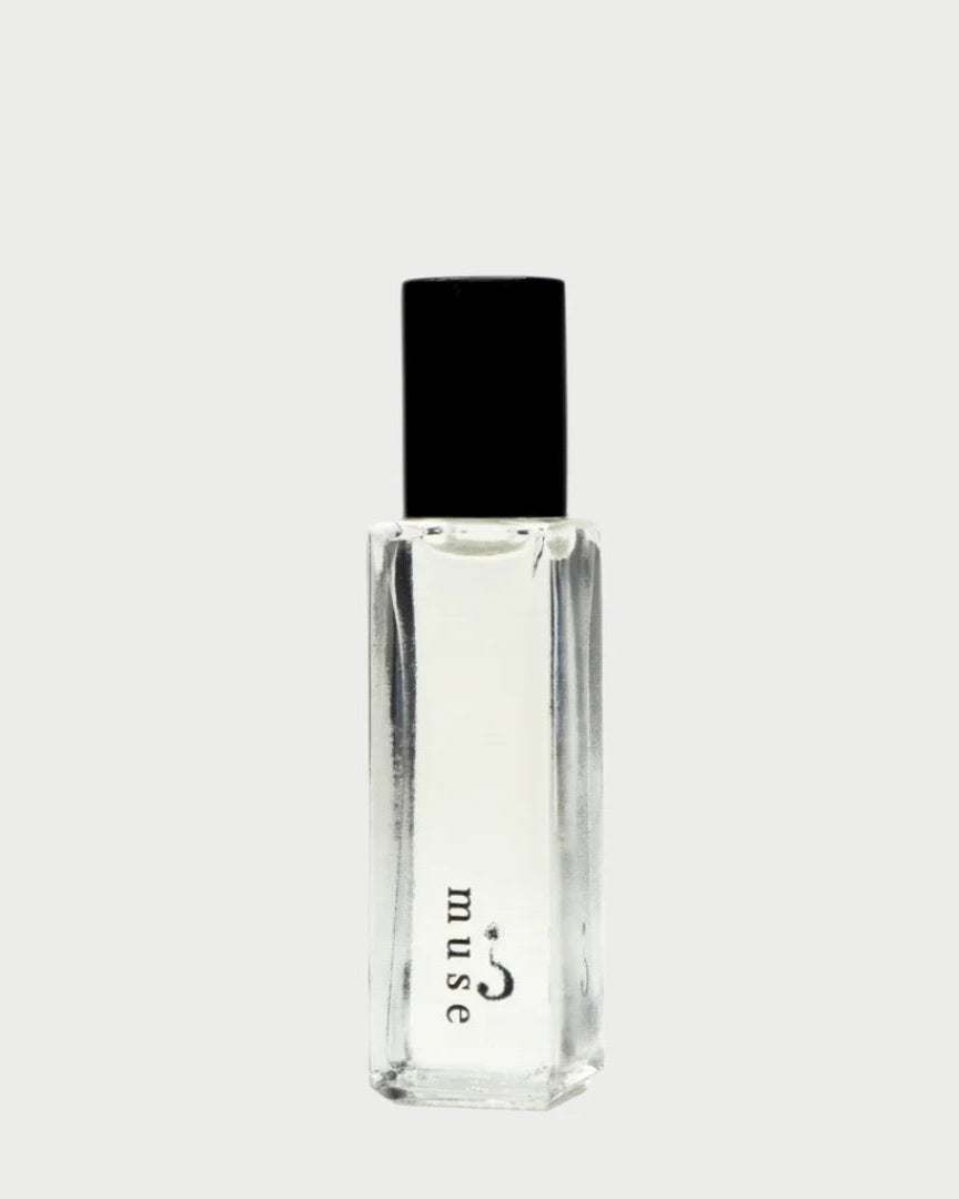 A clear glass bottle of Riddle Oil's Muse perfume, featuring a black cap, is centered on a plain light background. The word "muse" is printed vertically in lowercase letters, alluding to the sandalwood essence inside.