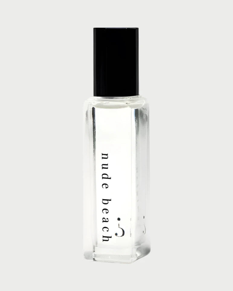A Riddle Oil - Nude Beach 8ml bottle, made of clear glass and sealed with a black cap, contains a transparent liquid. The label features the words "nude beach" in lowercase letters. The design is minimalist and sleek, showcasing the trending Nude Beach scent by Riddle Oil.