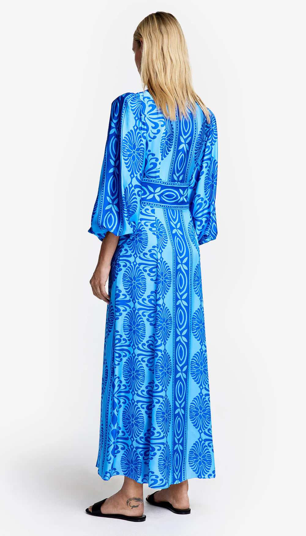 A blonde person wears a Smythe Crossover Midi Dress, long-sleeved with multi-blue hues and an intricate pattern, featuring a belted waist. They stand in black slip-on sandals, facing away from the camera against a white background.