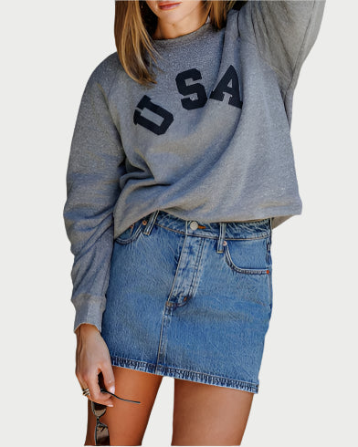 A woman in a heather grey Sunny Somewhere USA Raglan Crew and a blue denim mini skirt holds sunglasses. The image is cropped from chest to mid-thigh.