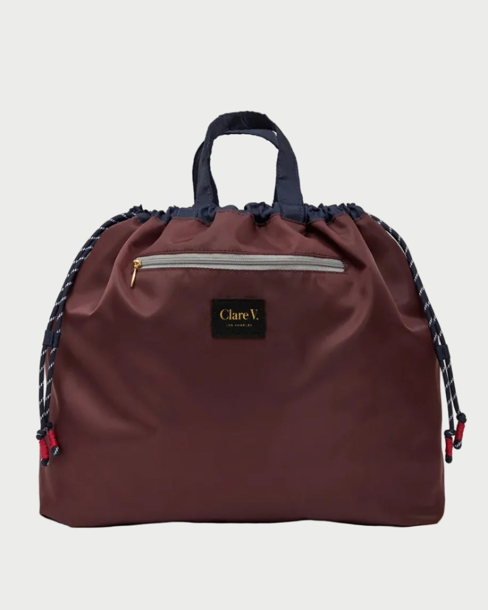 A dark burgundy drawstring bag with navy blue handles and braided side straps, featuring a front zip pocket and a black Clare V London brand patch.