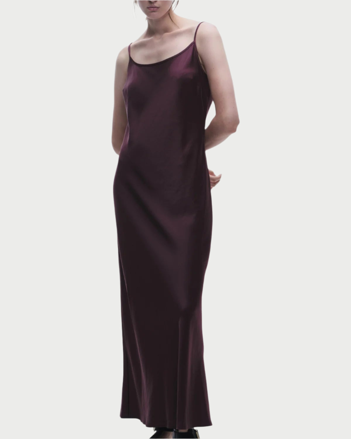 A person stands against a plain background, elegantly dressed in the Grape Haley Slip Dress by Saint Art NY, a sleeveless, dark purple satin gown with thin straps that gracefully cascades to the floor.