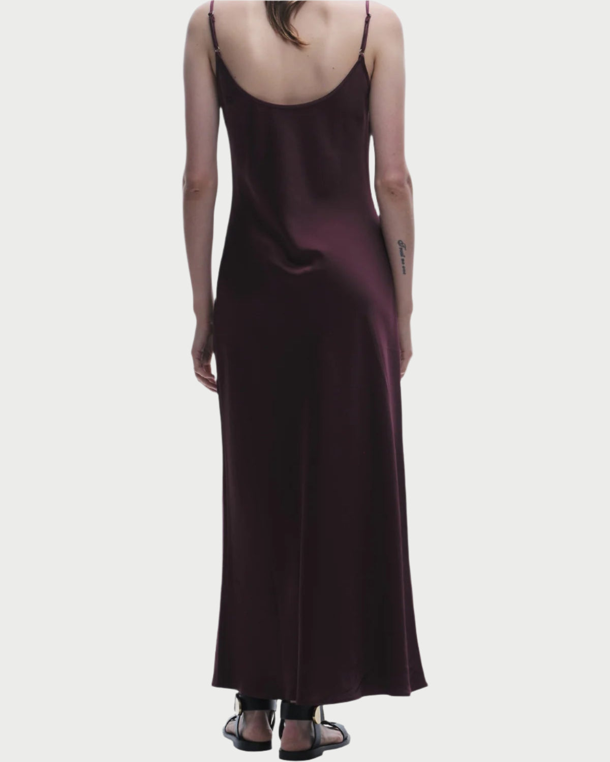 With grace, a person wears Saint Art NY's Grape Haley Slip Dress, a full-length satin piece with thin straps, beautifully contrasted with black sandals. Their long hair cascades down against the plain white backdrop—a true testament to Saint Art's craftsmanship.