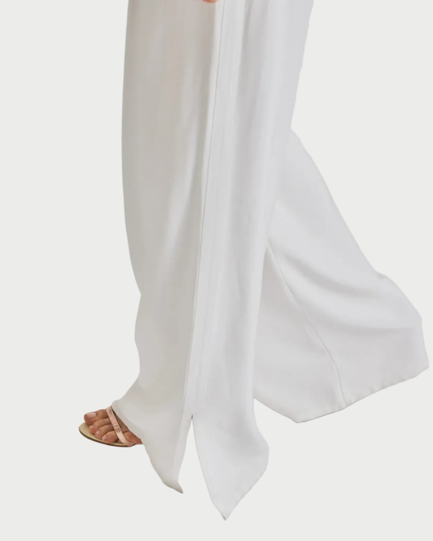 Wearing the Saint Art NY Neve Tuxedo Pant with a wide-leg silhouette and slit hem, paired with beige open-toe sandals, a model walks against a plain white background.