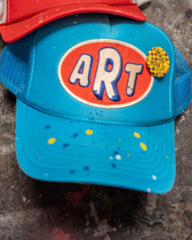 The Madeworn Art Trucker Hat is a limited-edition blue and orange cap with "ART" on the front, blue mesh sides, paint splatters, and a yellow "MAKE MORE ART!" pin on the right side.