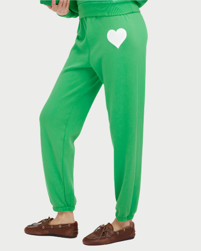 A person wears SPRWMN Heart sweatpants in bright green, featuring a white heart graphic on the back, paired with brown moccasin-style shoes for a fresh take on women’s joggers.