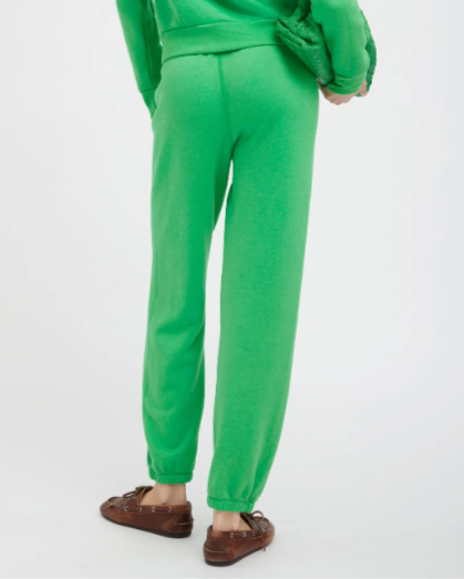 A person wearing SPRWMN Heart sweatpants in bright green with a matching sweatshirt stands facing away, holding a woven green bag under one arm. Brown loafers complete the look against a plain white background.