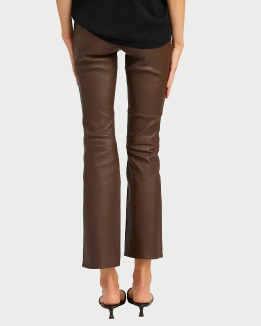 A person stands with their back to the camera, wearing SPRWMN 5 Pocket Ankle Flare pants by SPRWMN in French lambskin leather, styled with black heeled sandals and a chic black top. The high-waisted pants feature a subtle ankle flare.