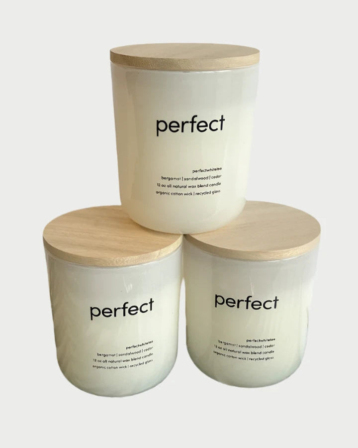 Three Perfect Candles from perfectwhitetee, each in white glass containers with wooden lids, are stacked in a pyramid. The candles are labeled "Perfect Candle" and beneath each label is a scent description highlighting bergamot, lavender, and cedar notes. They are elegantly displayed against a plain white background, featuring hints of sandalwood.