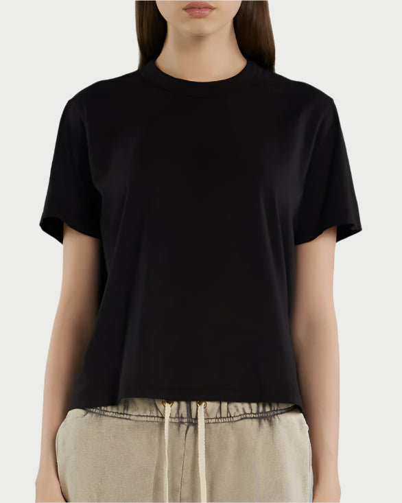 A woman wears the Les Tien Jane Classic Tee in plain black combed cotton with a relaxed fit, paired with light beige drawstring pants, shown from shoulders to hips against a light background.