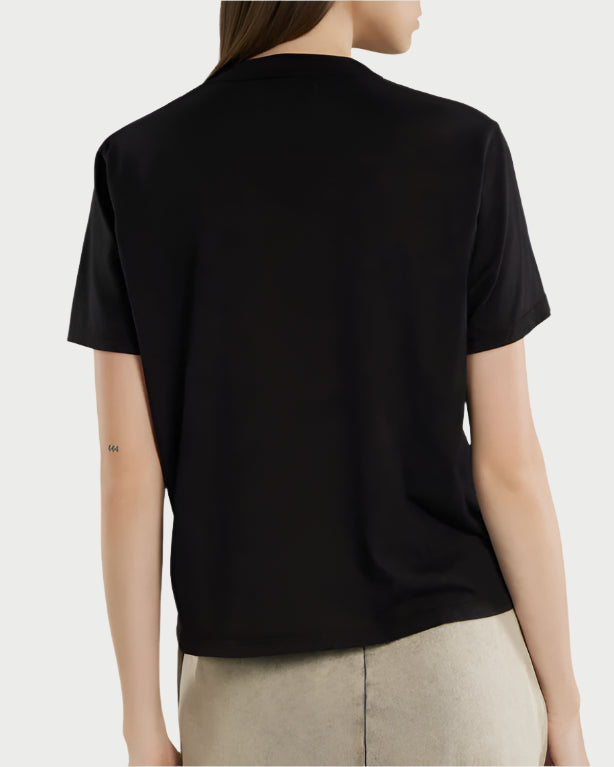 A person with long, straight brown hair, seen from behind, wears light pants and a relaxed fit black Les Tien Jane Classic Tee in combed cotton. A small tattoo is visible on their left arm.