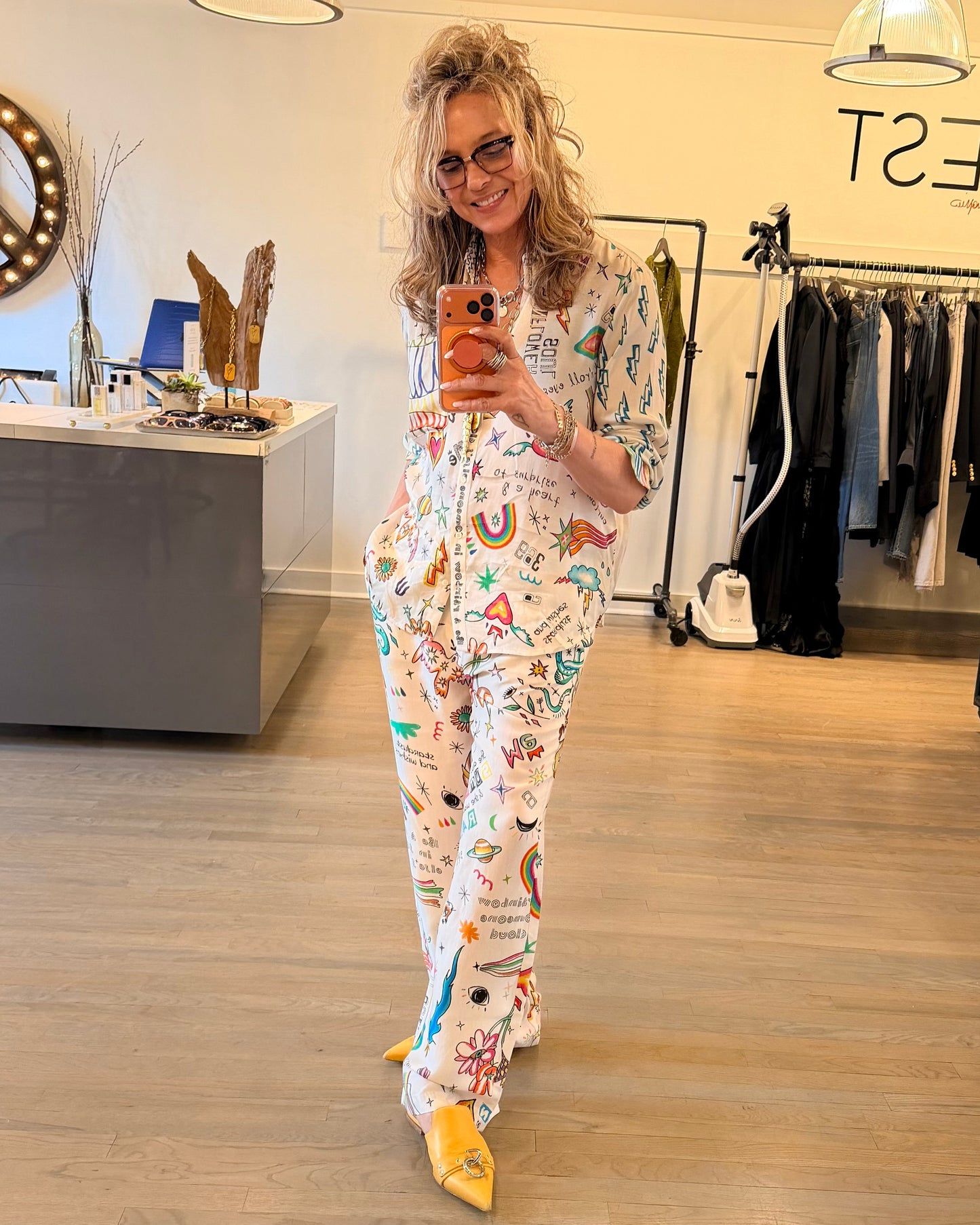 A woman takes a mirror selfie in a boutique, wearing glasses, yellow shoes, and the eye-catching ME369 Bailey Pants—a bold printed white piece by ME369. Clothing racks and a counter are seen in the background.