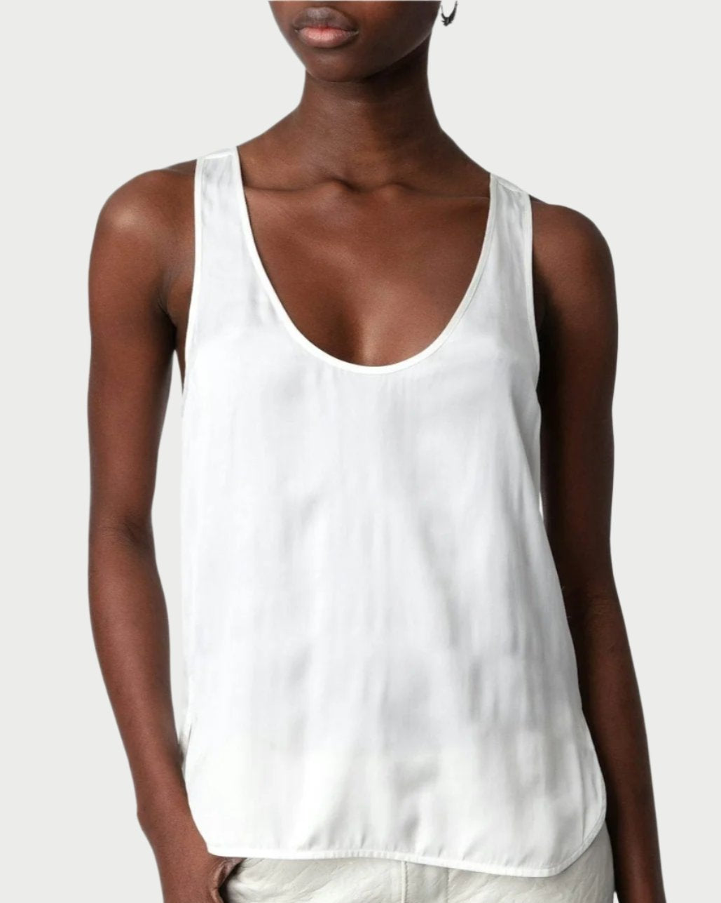 A person with close-cropped hair stands against a plain background, exuding a chic and sophisticated silhouette in black rectangular sunglasses, a Zadig & Voltaire Satin Cami, and light-colored pants. They have a confident expression and hold one hand in their pocket, showcasing versatile layering.