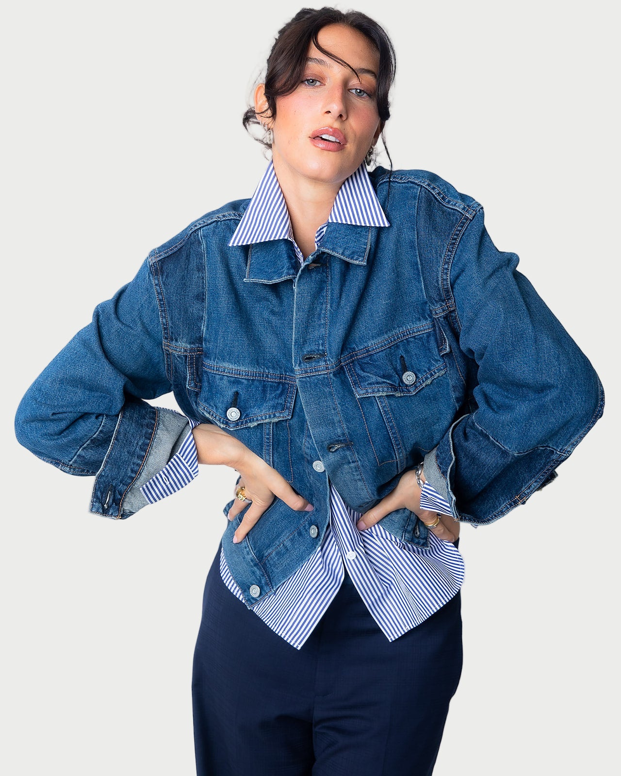 A woman with dark hair poses confidently in the Moussy Dover Two Tone Jean Jacket, layered over a blue-and-white striped shirt and dark pants, set against a plain light background.