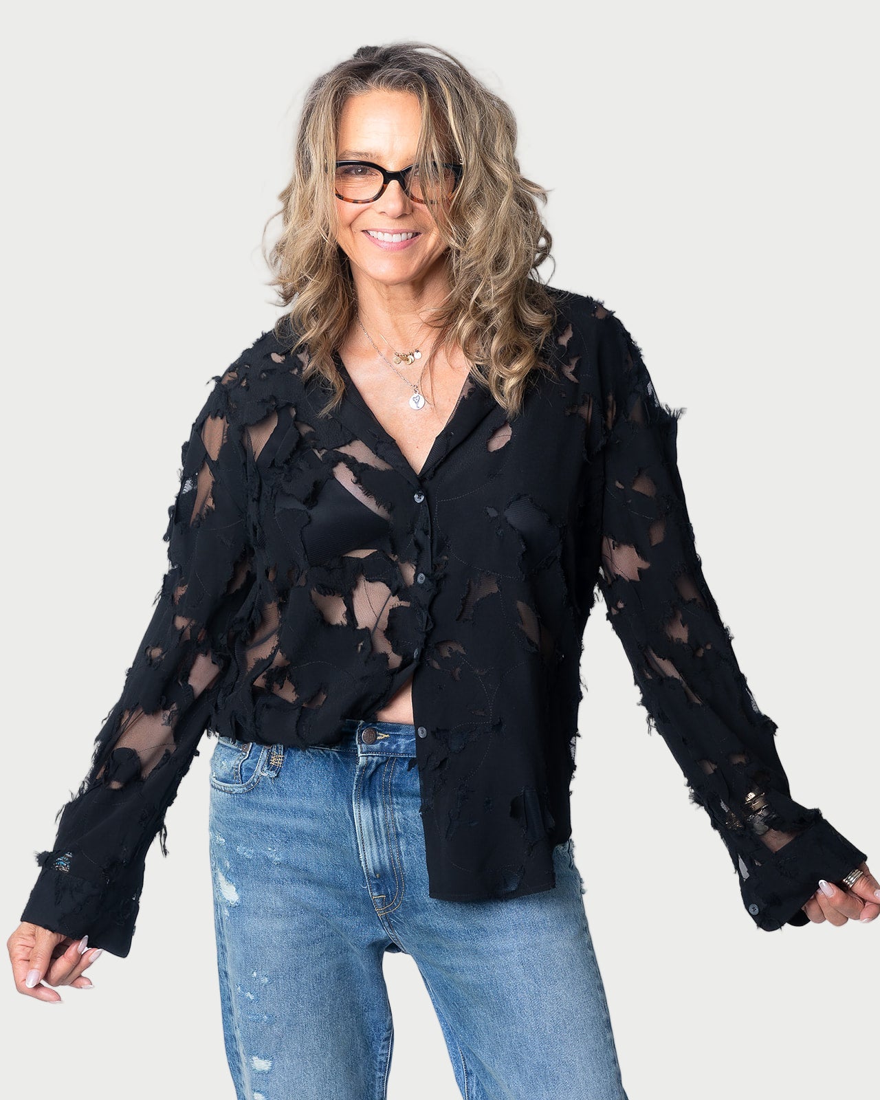 A woman with wavy blonde hair and glasses smiles at the camera, wearing a slim-fit R13 Slim Abstract Print Shirt in black, paired with blue jeans. The background is plain and light-colored.