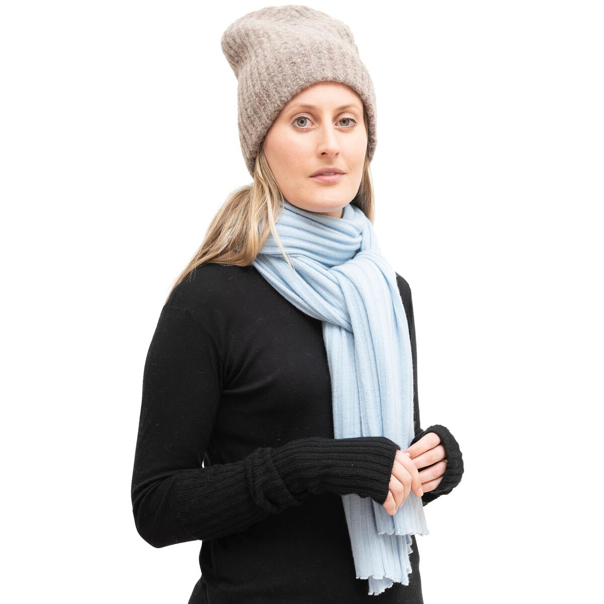 A person models added warmth in a Meg Cohen Alpaca Beanie, beige colored, with a light blue scarf wrapped around their neck, ends held in hand, and a black long-sleeve top against a plain white background.