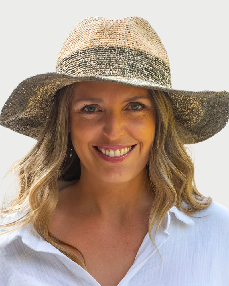 A smiling woman with long blonde hair wears the Travaux en Cours Raffia Floppy Hat and a white shirt, enjoying sun protection from the adjustable hat while standing outdoors by a blurred beach and ocean.