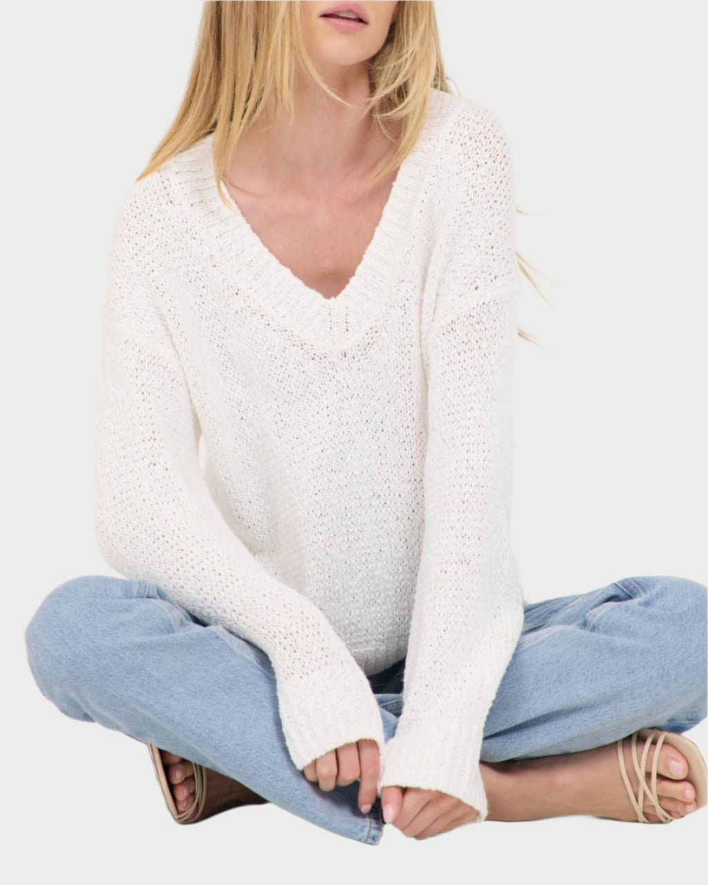 Sitting cross-legged against a white backdrop, a person wearing a Linnea V-Neck Sweater by One Grey Day and light blue jeans completes their casual look with flowing long blonde hair and sandals.