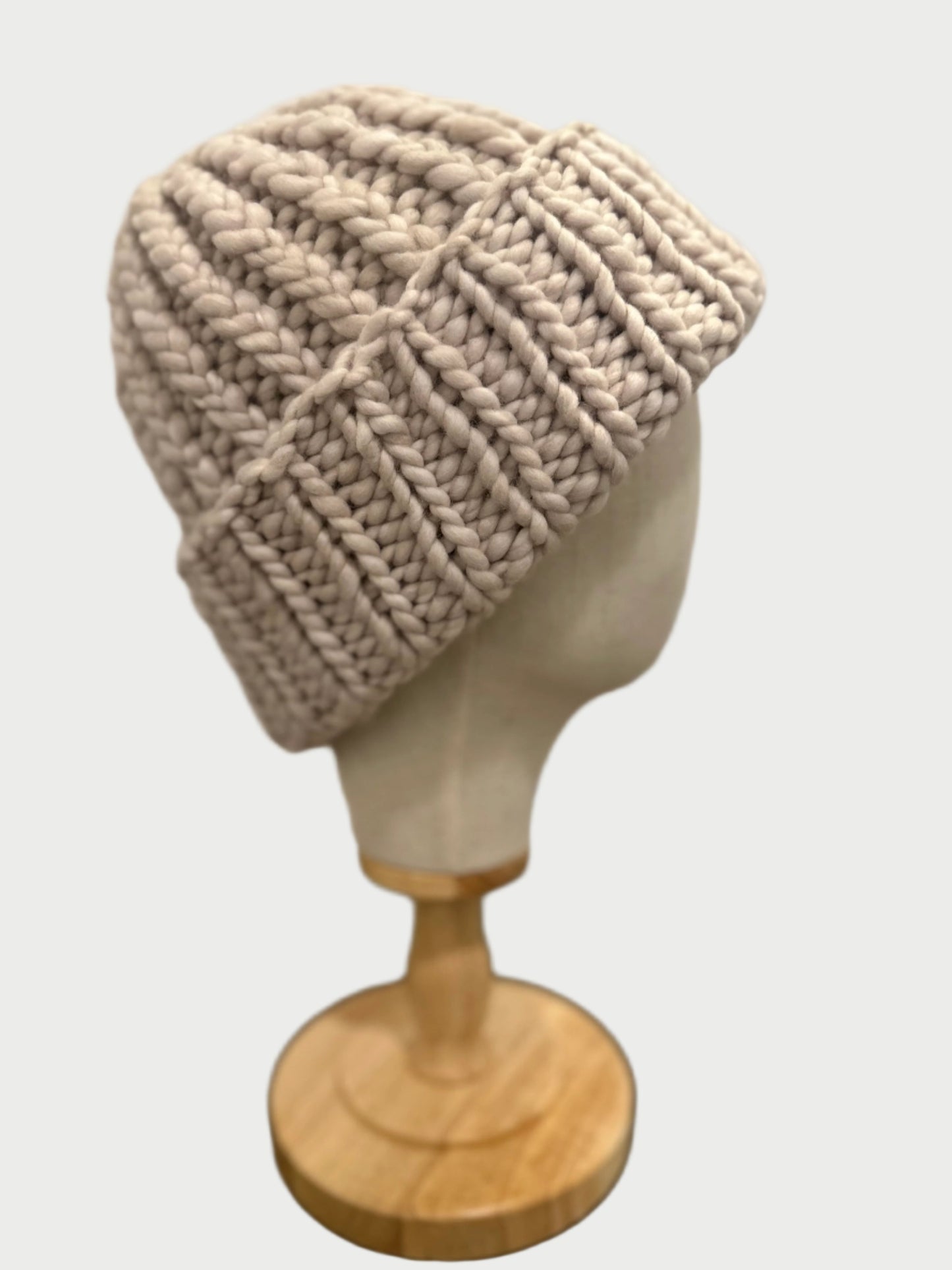 The Soft Wool Beanie by Meg Cohen, in beige and crafted from soft wool for warmth, is showcased on a wooden hat stand against a softly blurred background. This chunky knit beanie features a folded brim with thick ribbed stitching.