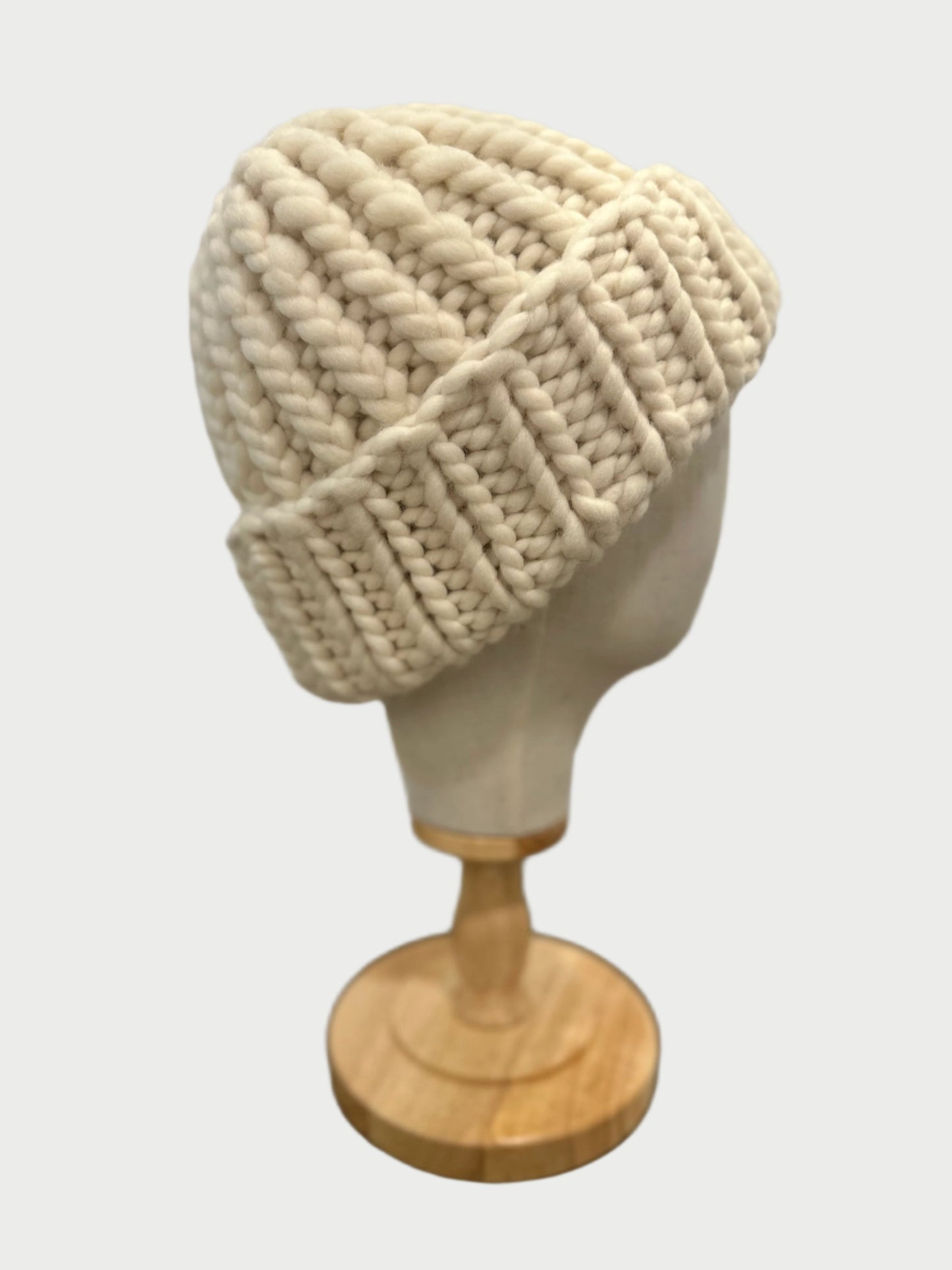 The Meg Cohen Soft Wool Beanie, in a cream-colored, chunky knit design and made from soft wool, ensures warmth while showcased on a wooden mannequin head. With its stylish folded brim, it rests gracefully on a dark wooden surface against a light gray backdrop.