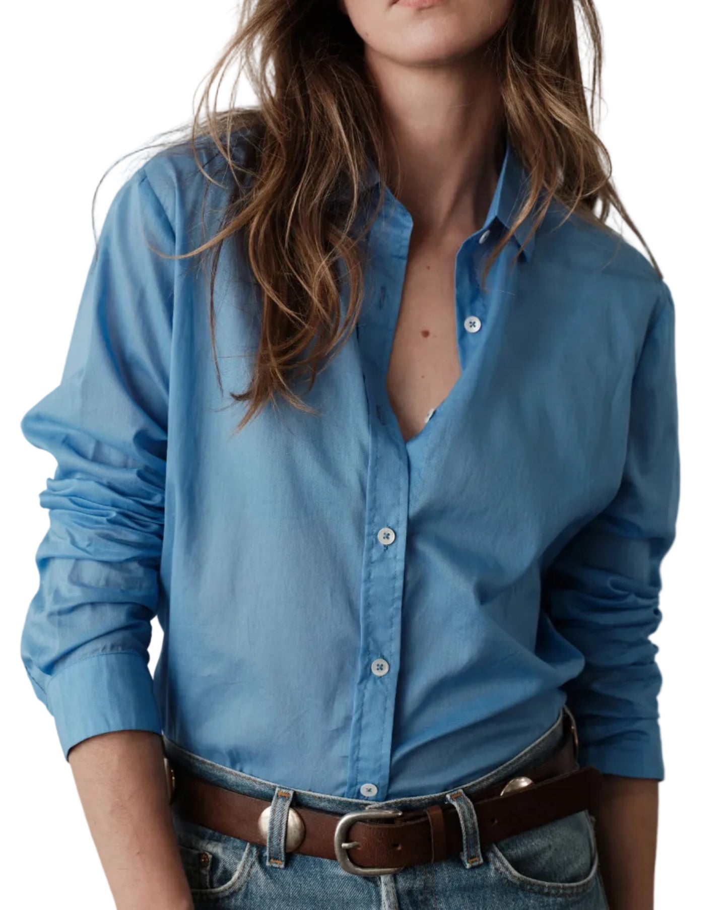 In Los Angeles, a woman wears the WEAR CISSA Classic Shirt in Cerulean Blue—loose-fitting, long-sleeved tissue cotton—tucked into blue jeans with a brown belt. Her wavy brown hair partially covers her face.