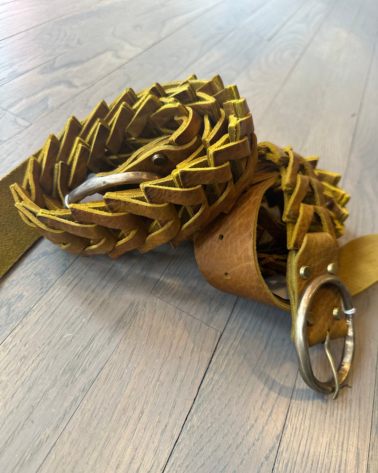 The Spike Leather Belt by Motif56, crafted in woven mustard yellow leather with a silver buckle and a triangle link design, is coiled on a light wooden floor, showcasing its versatile texture and depth.