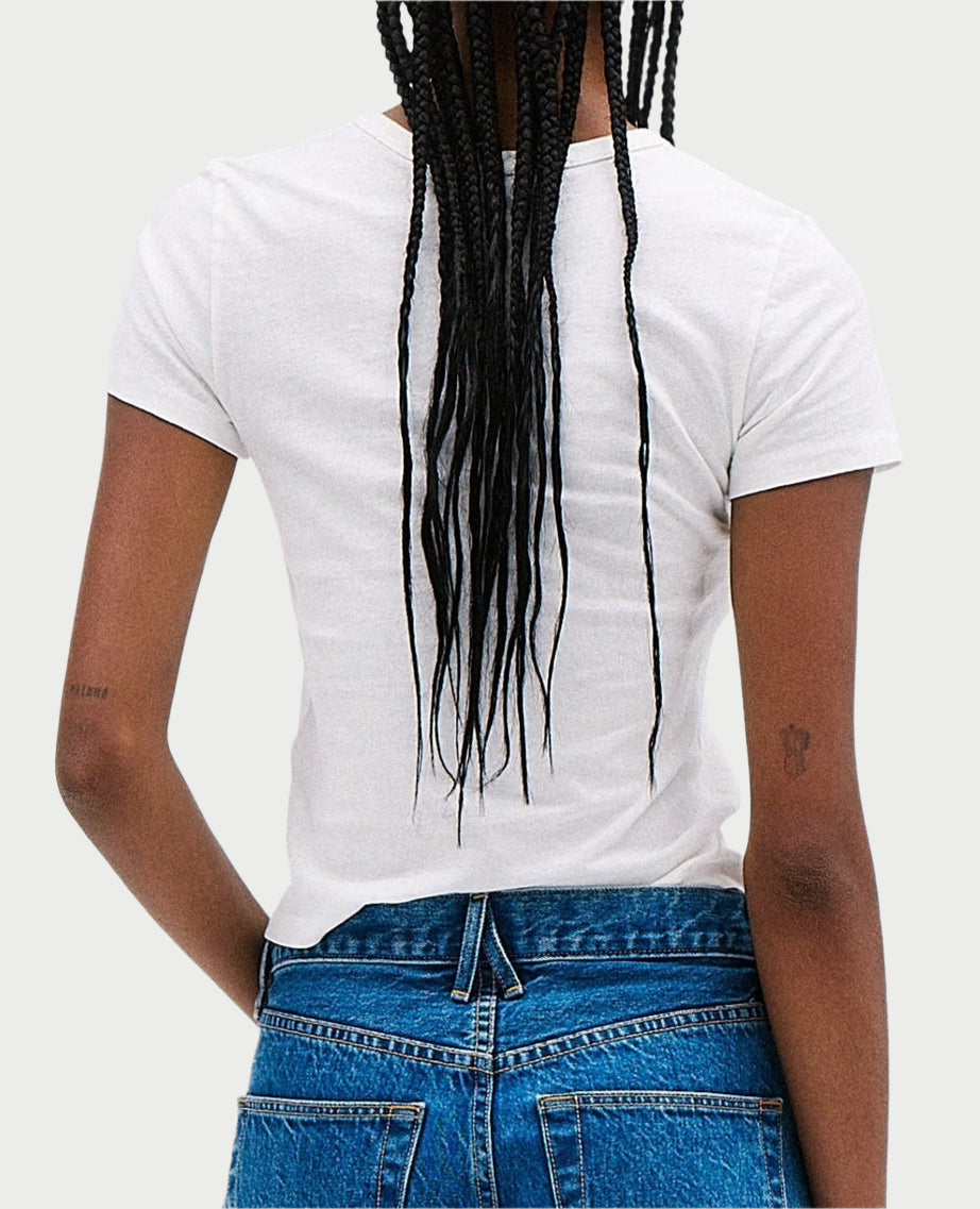 A person with long braided hair, wearing a white Easy Lightweight Tee and SLVRLAKE jeans, is viewed from the back. They have a small tattoo on their right arm, while the vintage look of the plain, light-colored background enhances the timeless style.