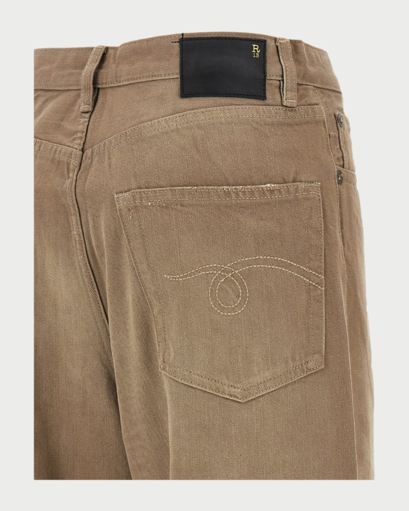 Close-up of the back pocket on R13 Damon Pleated Jeans in Sawdust, featuring beige stitching, unique looped embroidery, and a black leather patch with gold lettering at the waistband. Showcases an oversized slouchy fit.