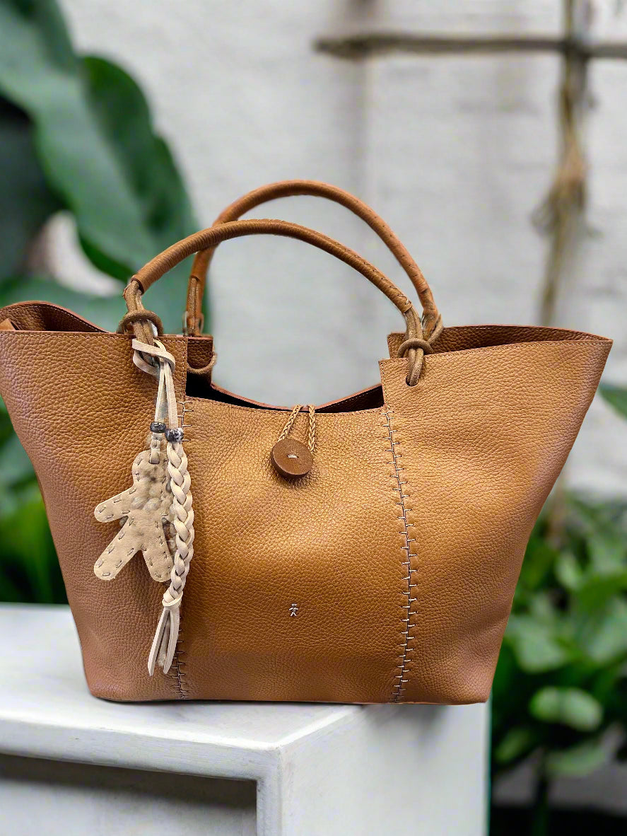 The Large Muflone Tote Bag by Henry Beguelin, crafted from tan calfskin leather in Italy with stitched detailing, features double handles, an adjustable shoulder strap, and a braided leather teddy bear keychain.