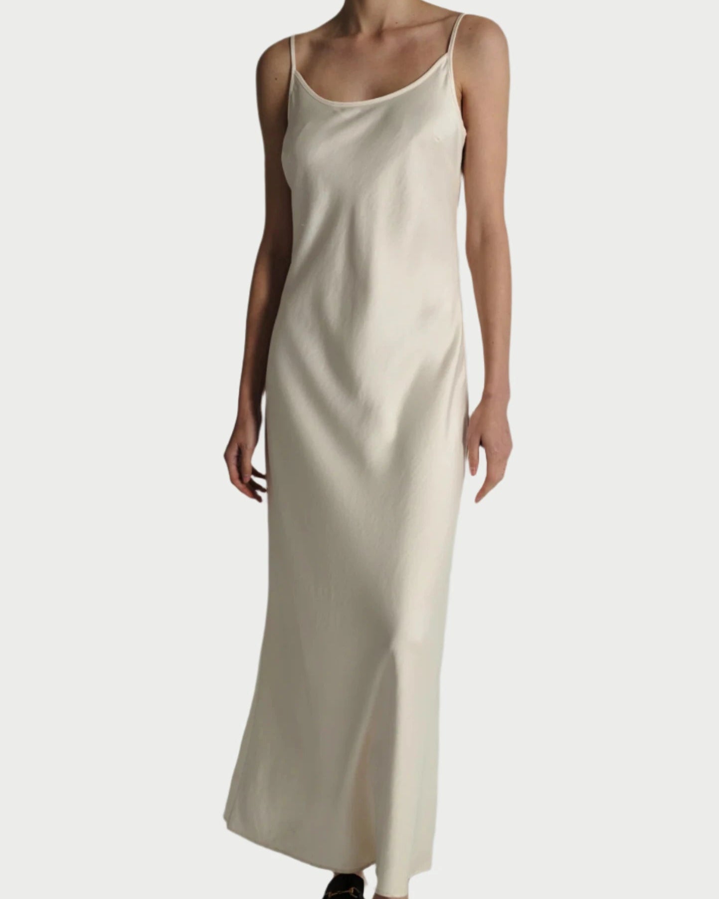 A person is wearing a long, sleeveless Saint Art NY Ivory Haley Slip Dress with spaghetti straps and black loafers. The dress has a simple, elegant design and appears to be made of silky, smooth, heavyweight charmeuse satin that is wrinkle-resistant. The background is plain and light-colored.
