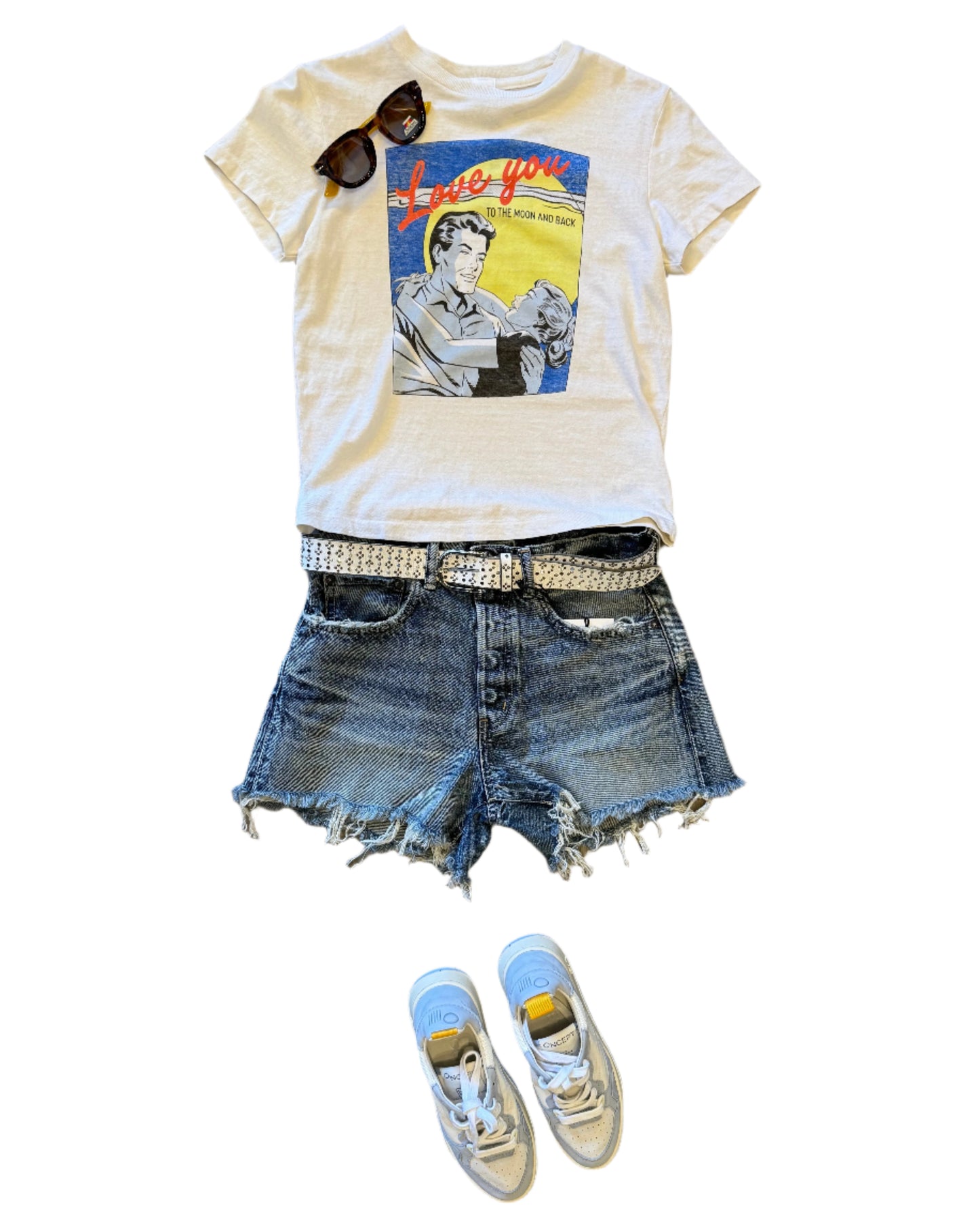 A casual outfit laid out on a wooden floor: a “Love you to the moon and back” graphic tee, Moussy Douglaston Denim Shorts with mid rise and mid blue wash, a studded belt, sunglasses, and white sneakers with blue and yellow accents.