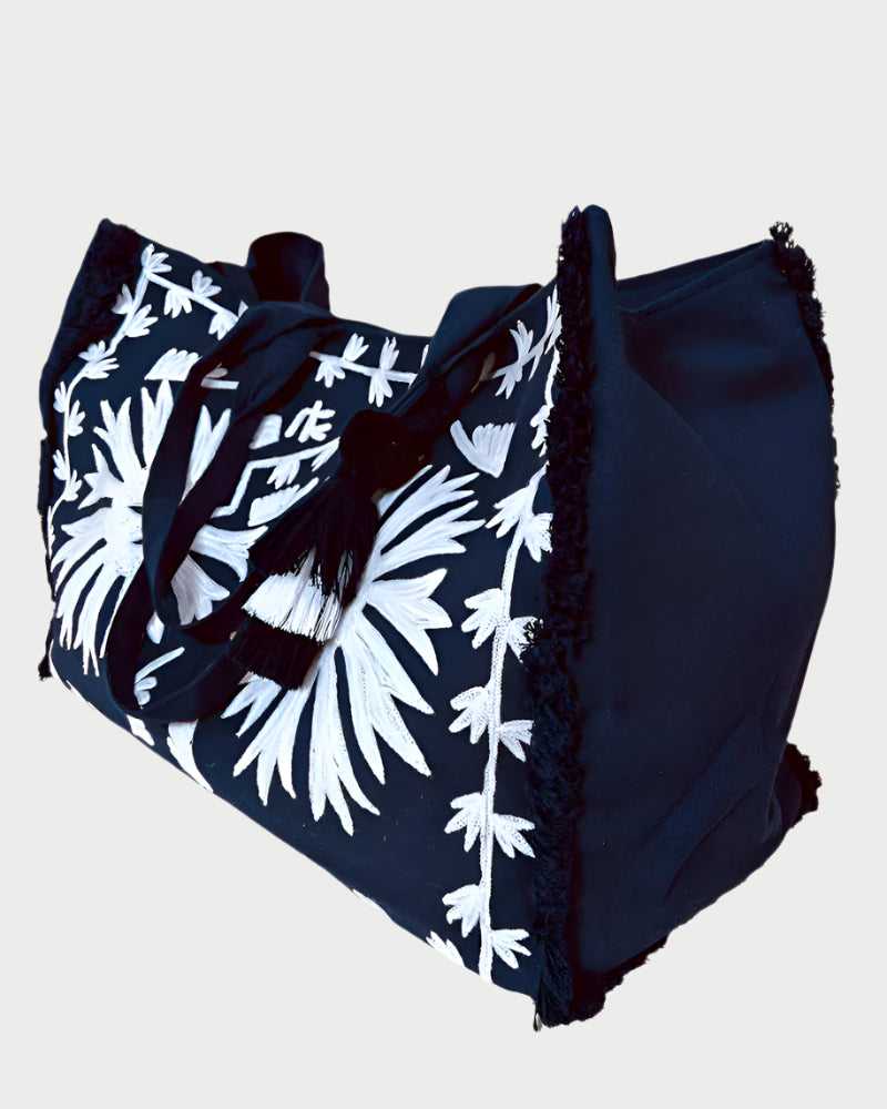 The DEBBIE KATZ Malan Beach Bag is a spacious black canvas tote with white floral embroidery, black handles, decorative fringe edges, soft textured fabric, and black-and-white tassel accents.
