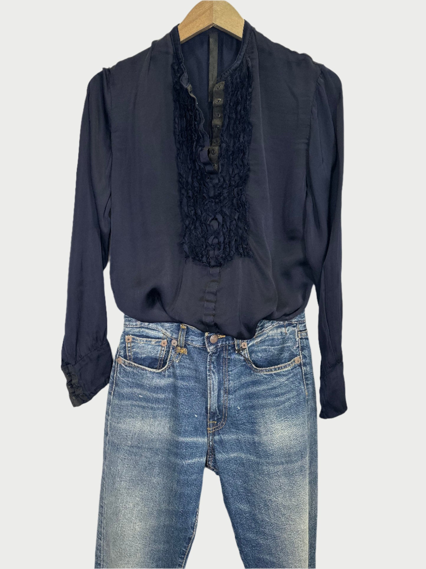 A long-sleeved, Wash & Wear Tuxedo Shirt with satin and ruffle details from Jaga LA graces the wooden hanger, exuding the flair of this Los Angeles designer. It pairs effortlessly with light blue, faded jeans for a chic look against the white background.