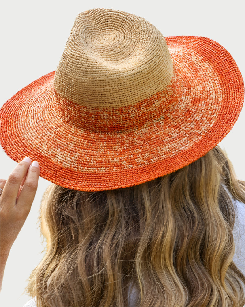 A person with long wavy blonde hair wears the Travaux en Cours Raffia Floppy Hat, featuring a tan crown and wide orange brim, holding the brim for sun protection by the water.