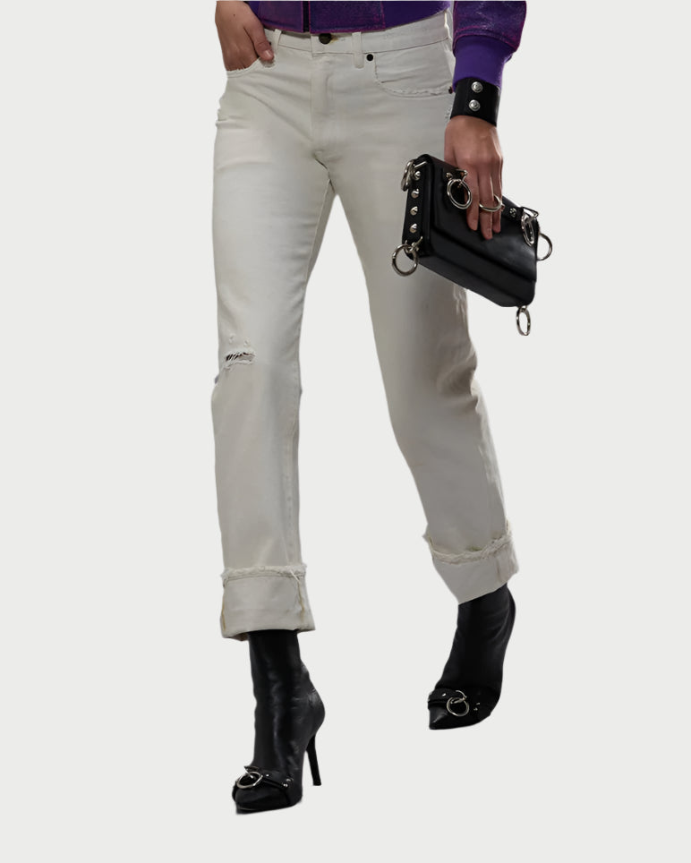 A person wears The Slim Boyfriend Jean in White Selvedge by R13, paired with black high-heeled boots, a purple top, and a black clutch with metal rings. The R13 jeans feature a small rip. Background is white.