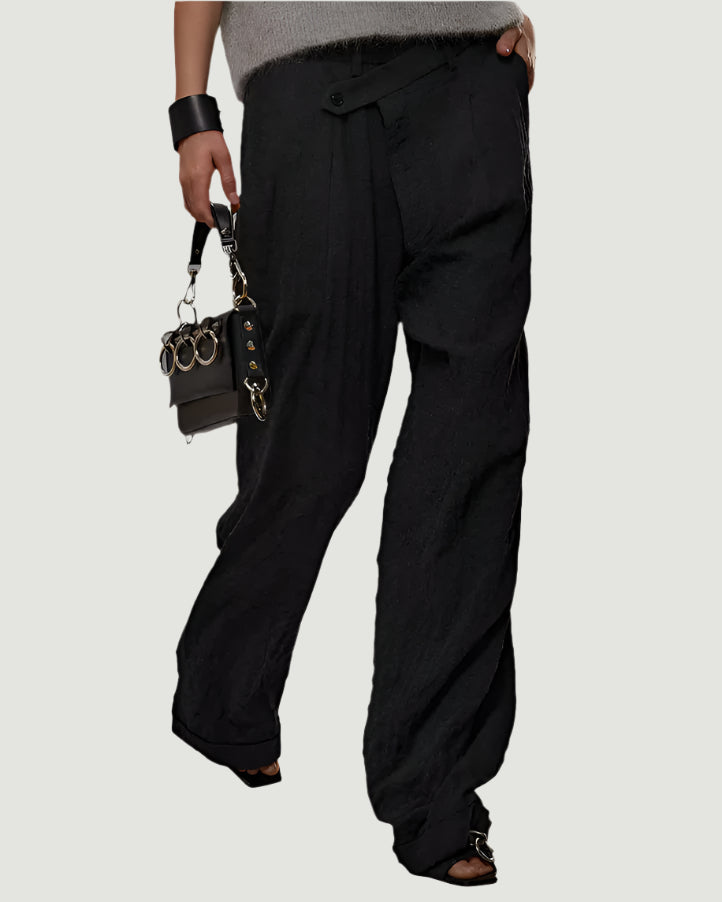 Woman wearing a sleeveless gray top and black pants holding a handbag against a plain background