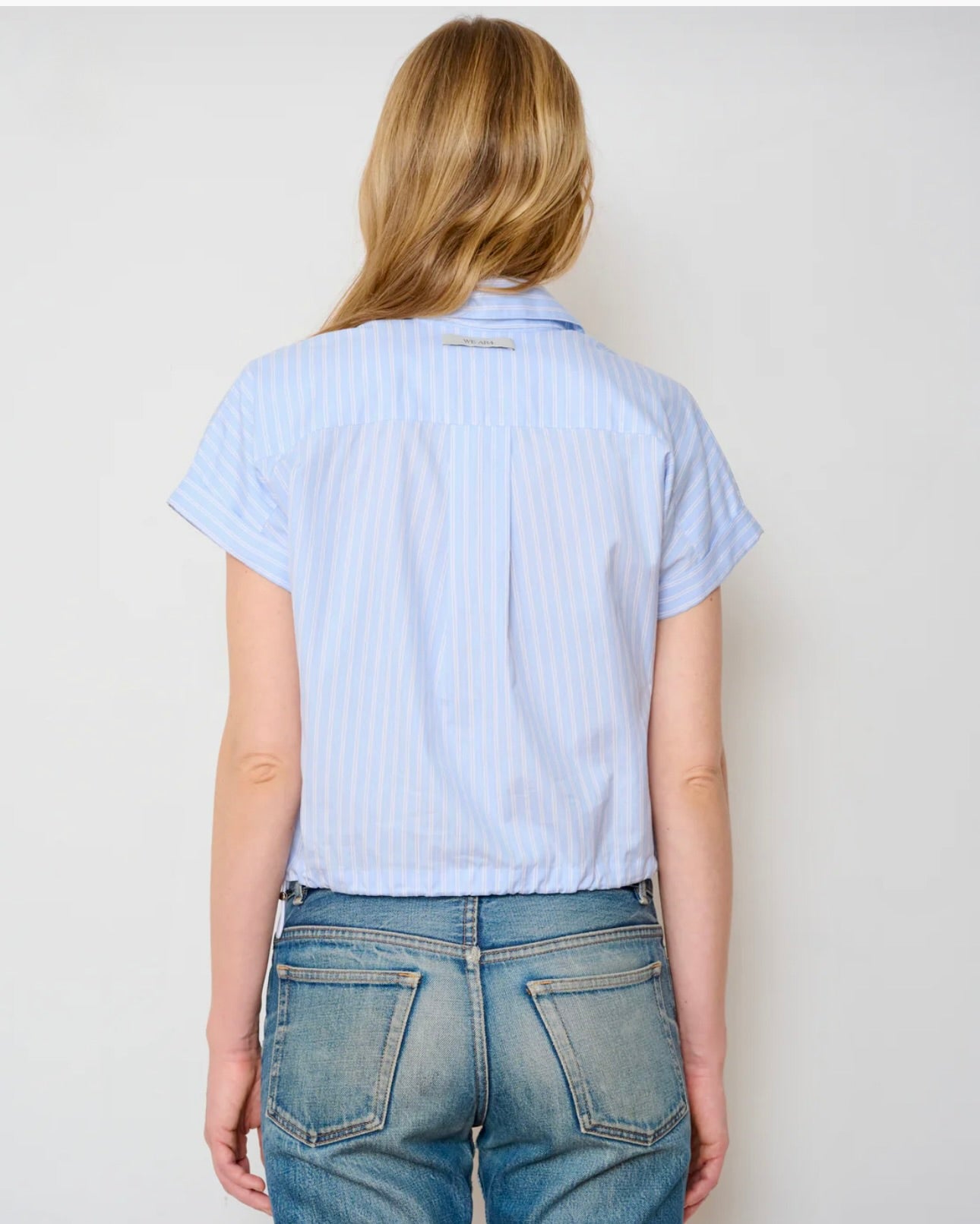 A person with long blonde hair is shown from the back, wearing the WE-AR4 Bungee Short Sleeve Shirt in light blue with thin white stripes and blue jeans. The shirt has a detachable collar and is made from upcycled cotton blend. Plain light background.