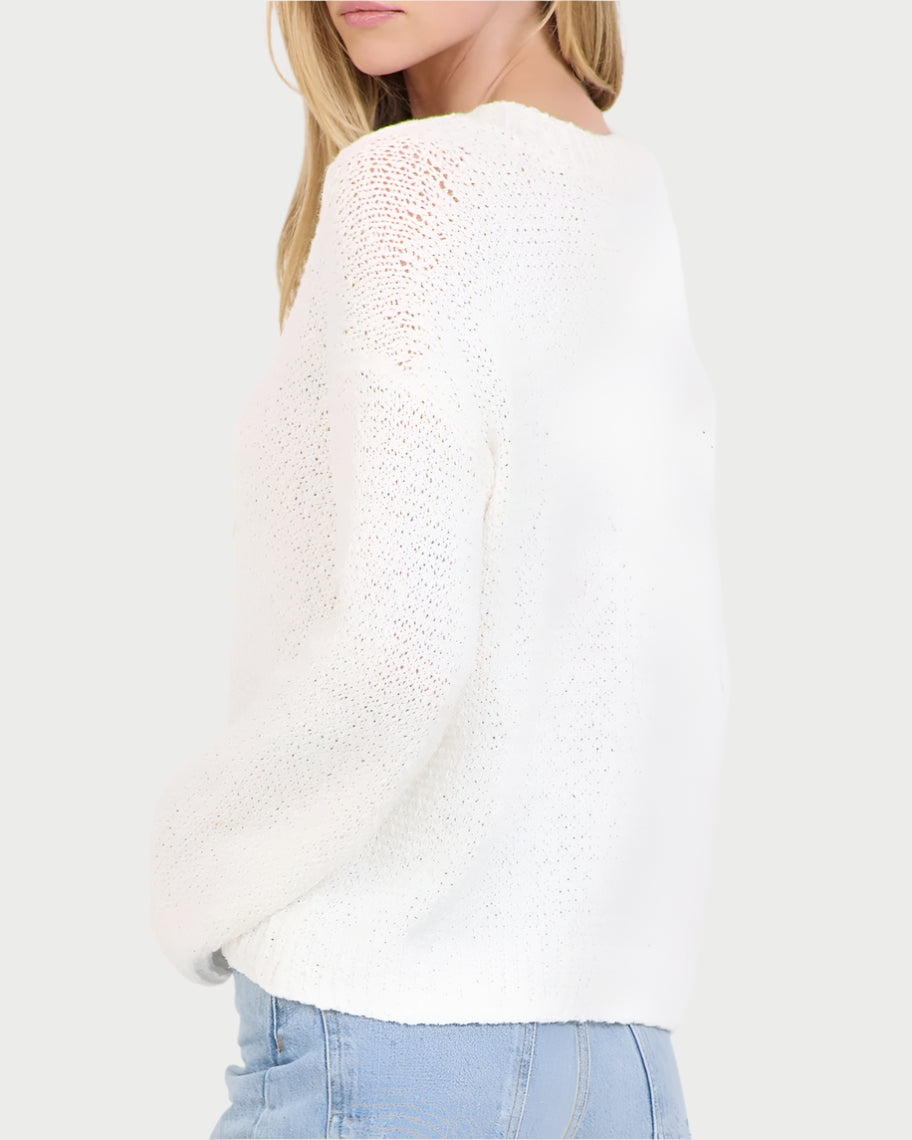 A person with long hair, angled sideways, wears a white Linnea V-Neck Sweater by One Grey Day and light blue jeans against a plain white backdrop. The textured chunky slub cotton of the sweater suits a relaxed style.